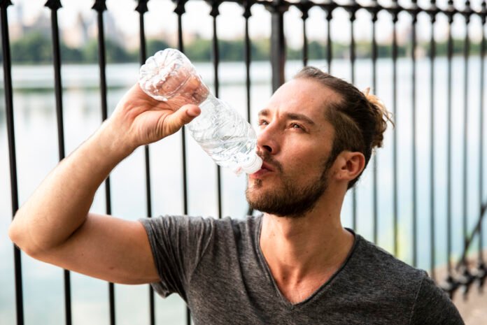 um jovem de barba bebendo agua mineral em uma garrafa.