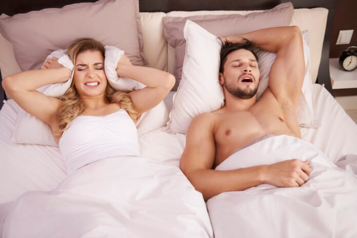 uma jovem tapando os ouvidos com o travesseiro devido ao ronco do seu marido ao lado.