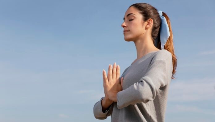 uma jovem em posição de yoga com as mãos postas num fundo azul claro.