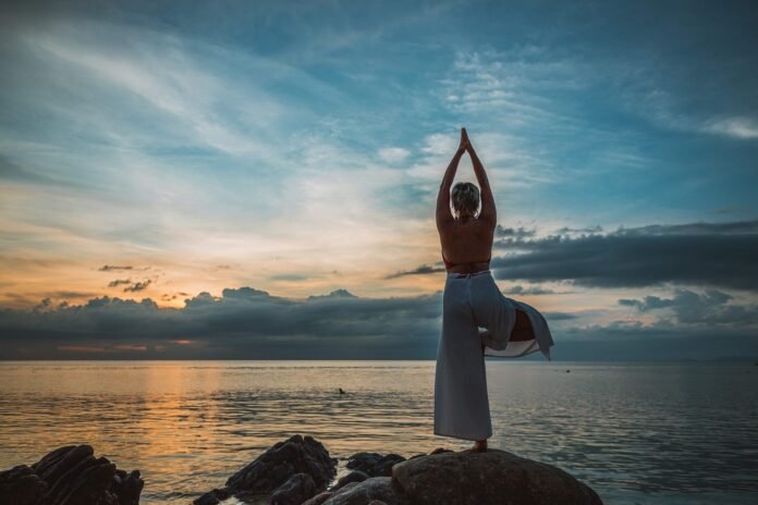 uma mulher vista pelas costas em posição de yoga diante de um lindo por do sol m um lago.