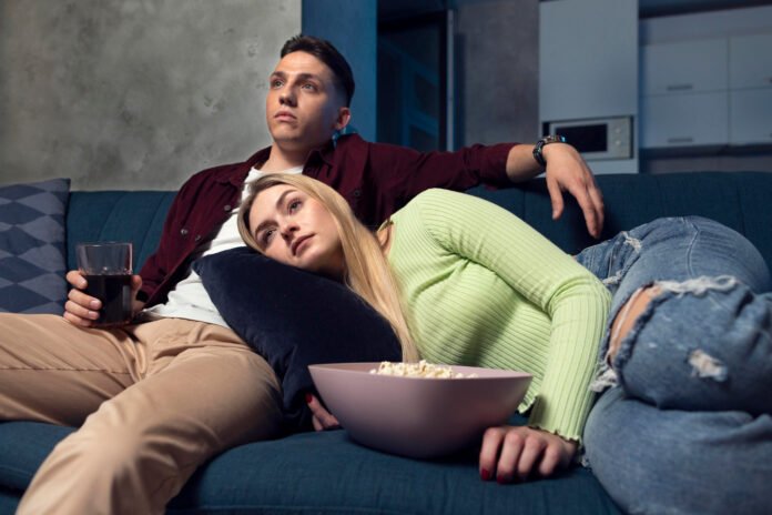 um casal no sofá, ele sentado e ela deitada no colo dele, assistindo televisão e comendo pipoca.