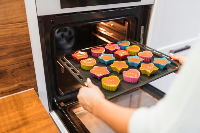 mãos colocando uma bandeja contendo formas de silicone com alimentos no forno.