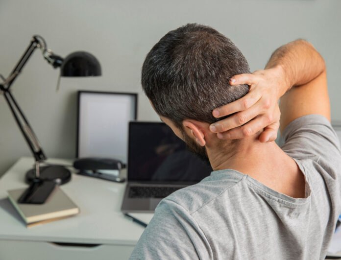um homem jovem de costas, sentado numa cadeira de trabalho, massageando a nuca.