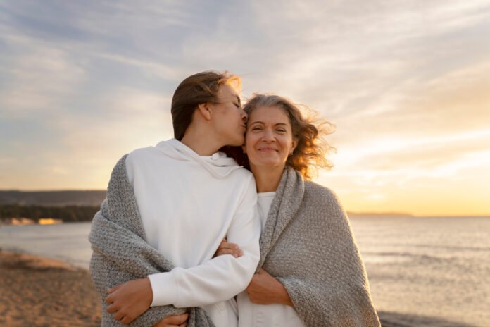 imagem de uma mae e um filho adulto, abraçados em uma praia ao entardecer.