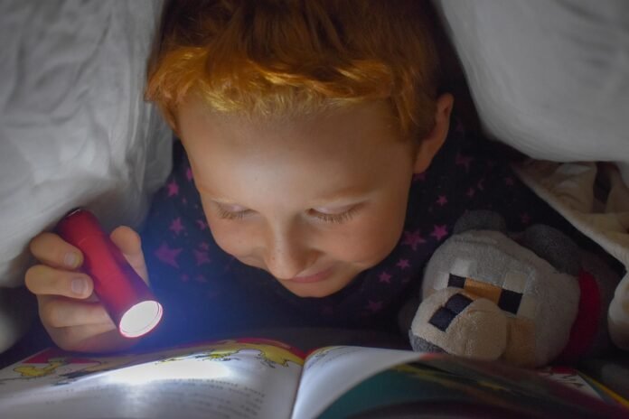 imagem de uma criança lendo um livro, debaixo do lençol, com uma lantera.