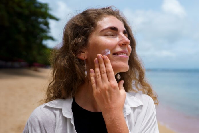 Uma jovem mulher aplicando bloqueador solar no rosto, em uma praia.