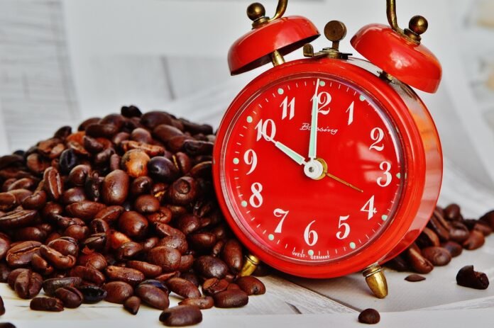 grãos de café sobre uma mesa branca, com um relógio despertador vermelho.